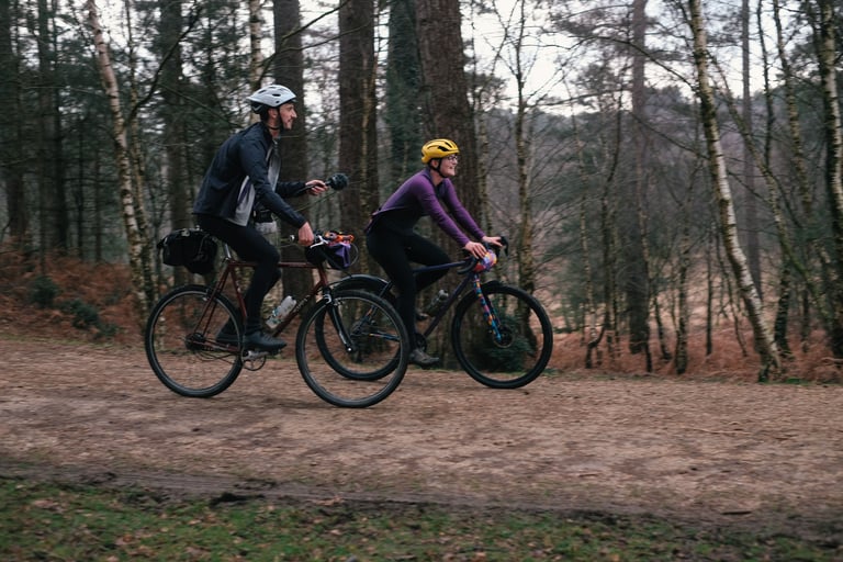 Unpaved Podcast hosts Katherine and Tom ride a gravel track through woodland, one holding a microphone.