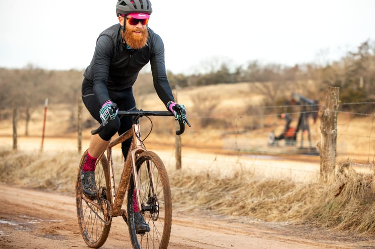 Rider on the Salsa Stormchaser on a muddy red Oklahoma gravel road, with an oil pumpjack behind a fence.