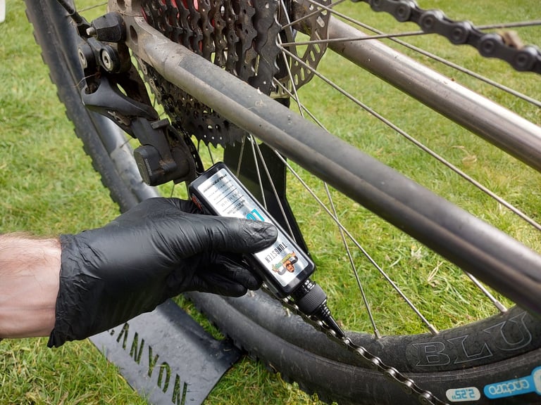 Gloved hand dripping Tru-Tension BananaSlip wet lube onto a mountain bike chain beside the cassette.