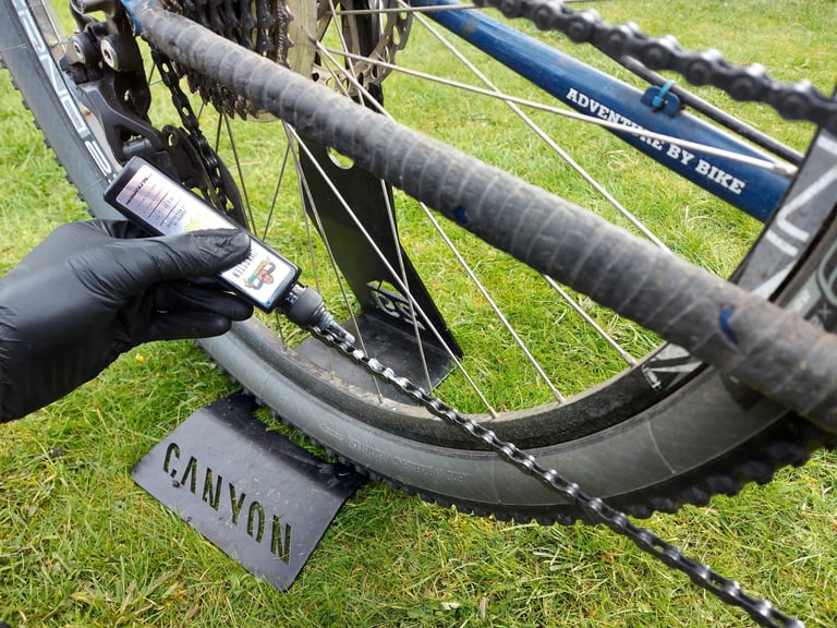 Applying Tru-Tension BananaSlip all-weather lube to a bike chain, gloved hand holding the bottle.