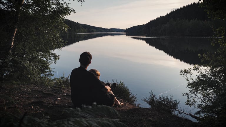 Father and son sit at the edge of a still lake at dusk, wooded hills beyond, at the end of a day's bikepacking.