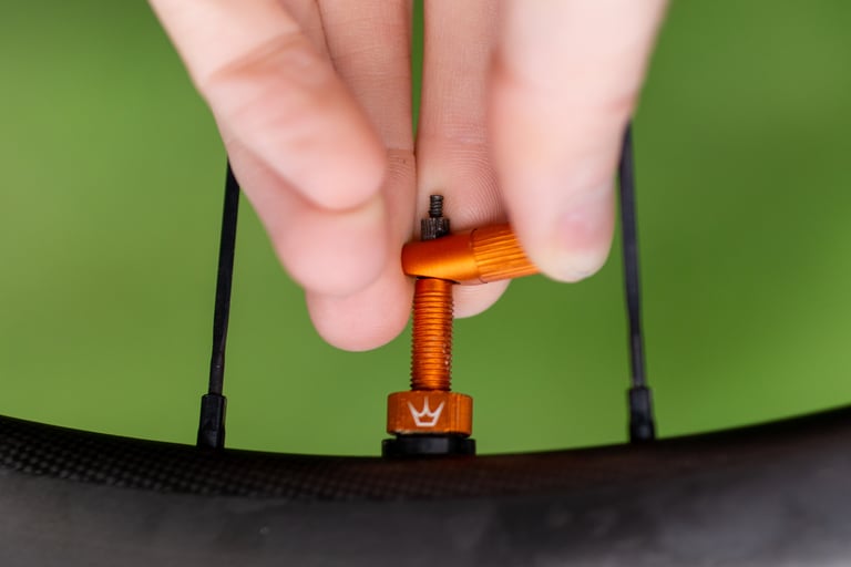 Fingers using the Peaty's tubeless valve cap as a valve core remover on an orange valve fitted to a carbon rim.