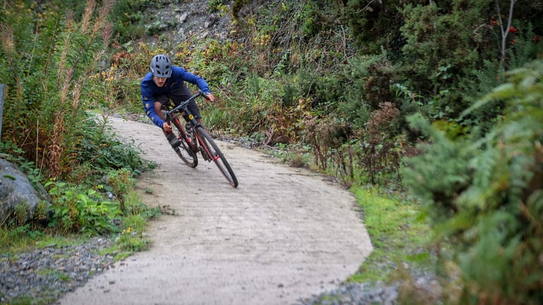 Rider leans a FUSTLE Causeway GR1 into a bend on a hard-packed gravel trail cut through scrubby hillside.