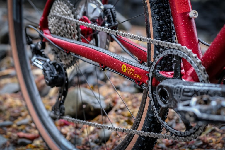 Close-up of the red FUSTLE Causeway GR1 chainstay, Race Face crank, rear mech and tan-wall tyre on wet pebbles.