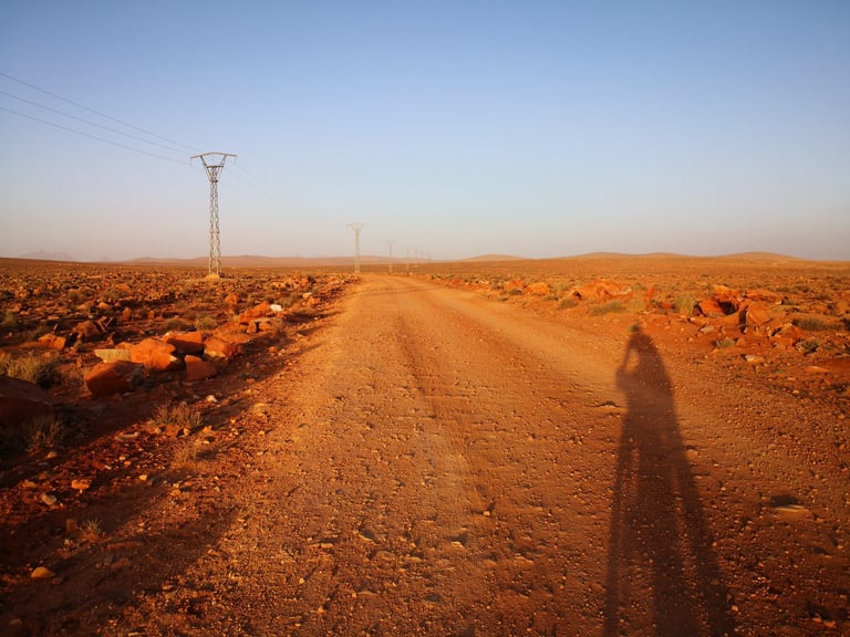 Rider's long shadow falls across a red dirt road lined with pylons at dusk on the Atlas Mountain Race, near Tafraoute.