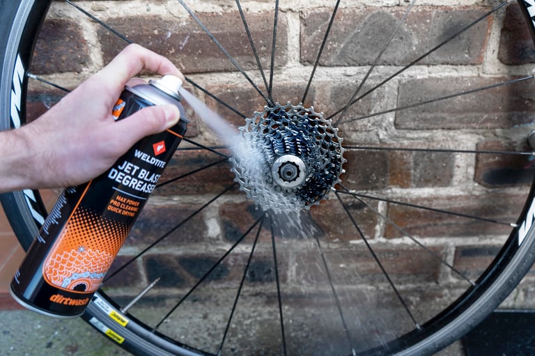 Weldtite Jet Blast Degreaser sprayed at a road bike cassette on a wheel propped against a brick wall.