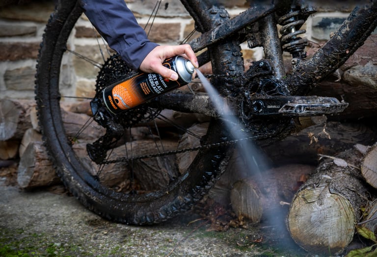 Jet Blast degreaser sprayed at the chain and chainring of a mud-caked full-suspension mountain bike.
