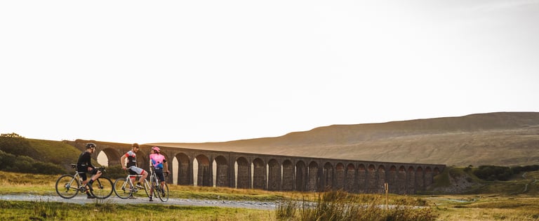 Three riders stop to talk on their bikes in front of Ribblehead Viaduct in North Yorkshire at sunset.