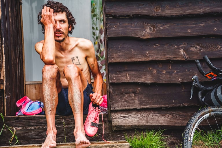 Exhausted Lachlan Morton sits barefoot holding a pink shoe at GBDURO checkpoint 1, his bike beside him.