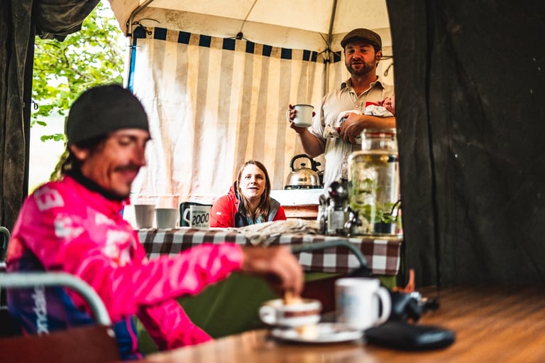 Lachlan Morton in a pink jersey eats breakfast in a tent at GBDURO checkpoint 1, a man holding a baby behind him.