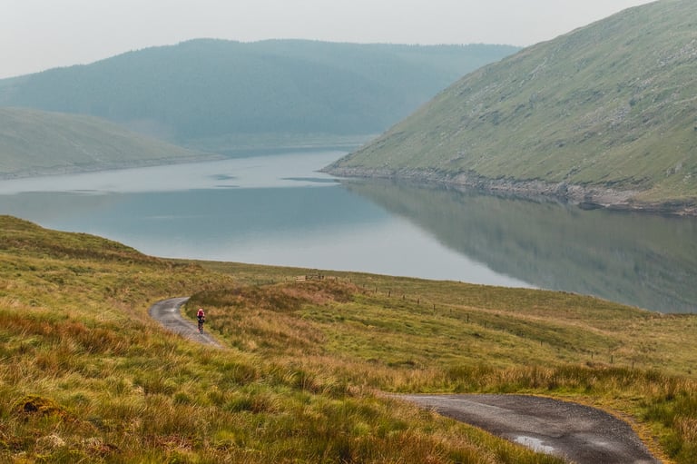 Lachlan Morton rides alone up a narrow road above a lake between bare hills on stage 2 of the GBDURO.
