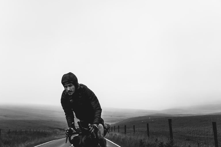 Lachlan Morton in a hooded jacket rides a misty empty road in black and white during the GBDURO.
