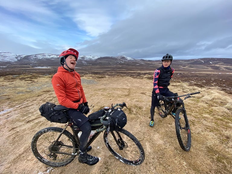 Two bikepackers grin on a moorland track in the Cairngorms, one astride a loaded gravel bike, snowy hills behind.