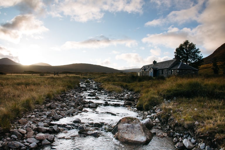 Ben Alder Cottage, a remote stone bothy beside a rocky stream with hills behind in low evening light.