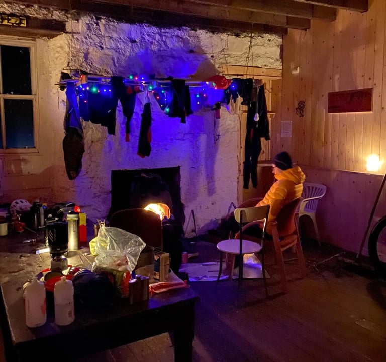 Inside Glas-allt-Shiel bothy, kit hangs to dry above the fire while a rider in a yellow jacket sits beside it.
