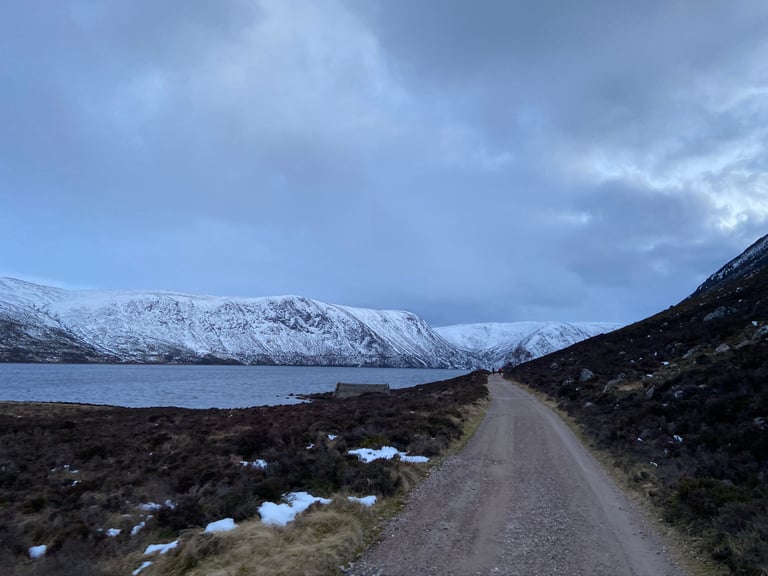 Gravel track beside Loch Muick in fading light, with snow-dusted hills rising behind the water.