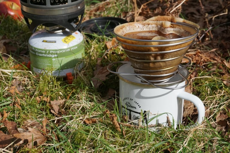 Camping stove and collapsible dripper over an enamel mug on grass, brewing a mid-ride coffee outdoors.