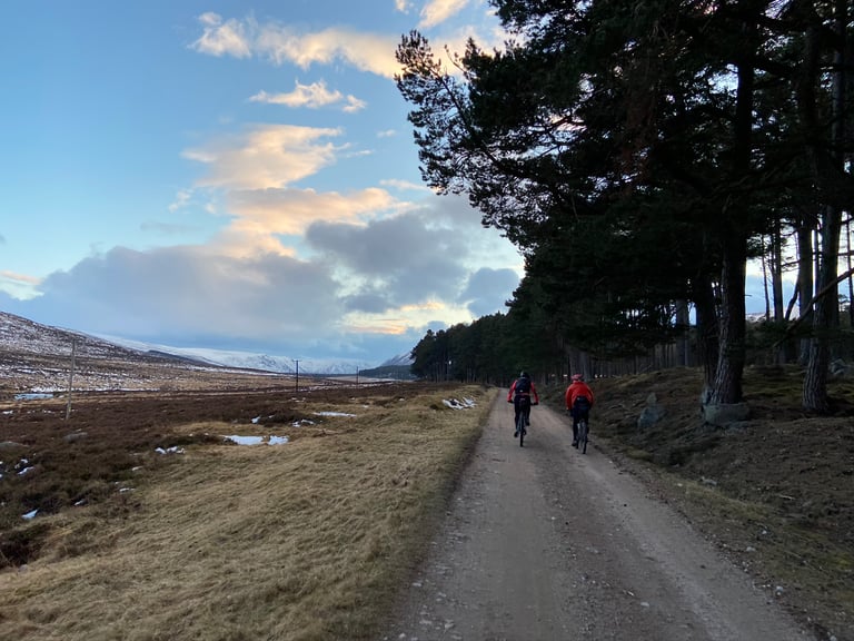 Two riders on a gravel track beside heather moorland at dusk, snowy hills ahead, on the Cairngorms route.