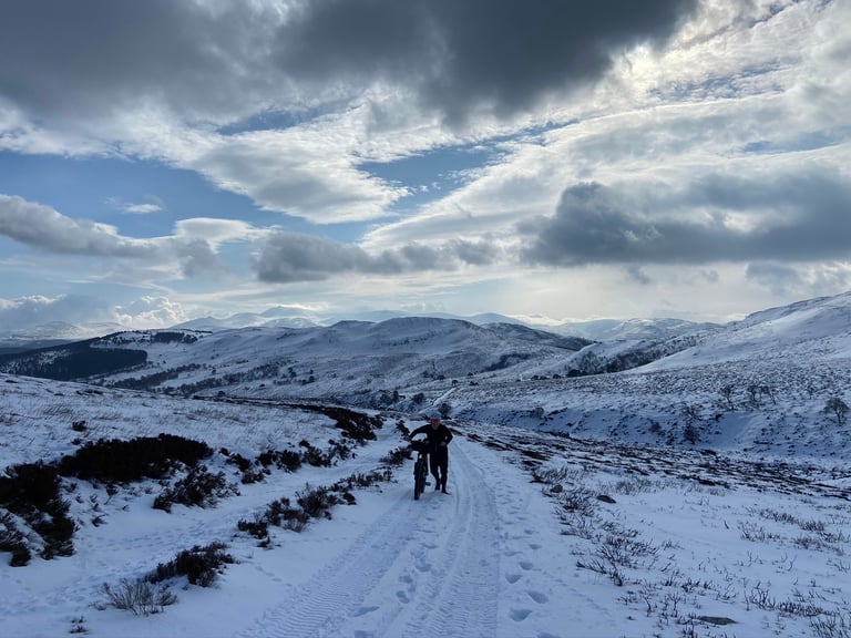 Rider pushes a loaded bike along a snow-covered track in a wide Cairngorms valley under broken cloud.