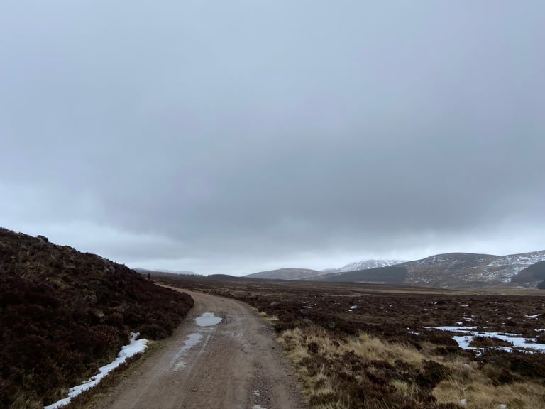 Gravel track curving across heather moorland in the Cairngorms under grey cloud, with snow patches and hills ahead.