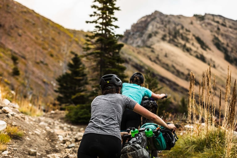 Two bikepackers push loaded bikes up a rocky mountain track in the Canadian Rockies.