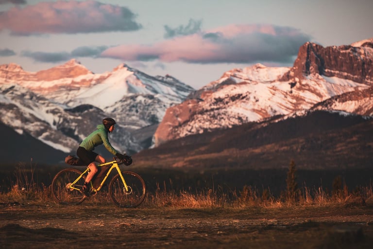 Sarah Hornby rides a yellow bikepacking bike on a track below snow-capped Canadian Rockies peaks in low sun.