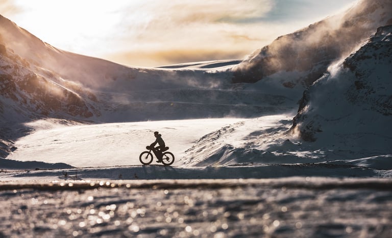 Bikepacker rides a loaded bike across wind-blown snow between mountain slopes in the Canadian Rockies.