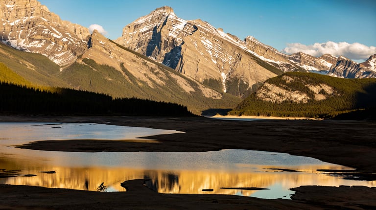 Tiny silhouette of a bikepacker riding beside a reflective lake below Rocky Mountain peaks in Alberta.