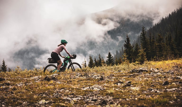 Bikepacker on a teal bike rides rocky alpine ground below low cloud and forested slopes in the Canadian Rockies.