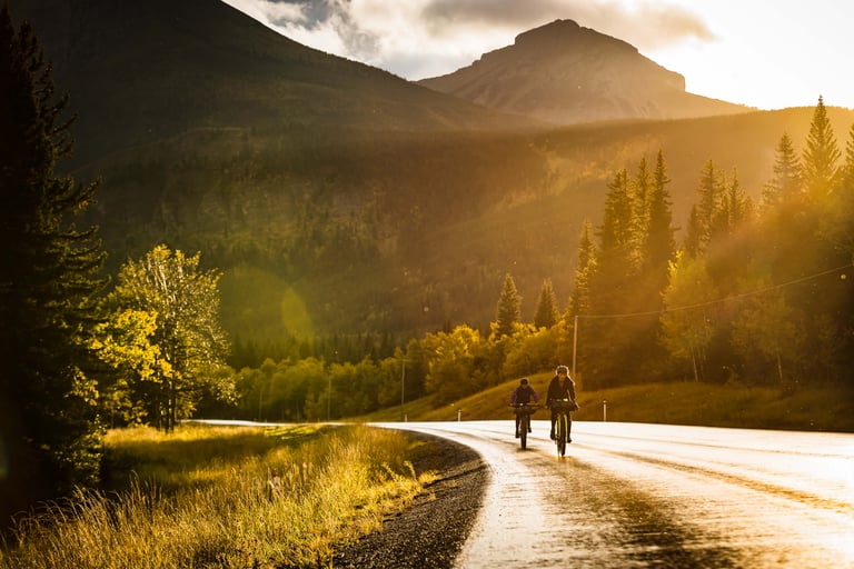 Two bikepackers ride a wet, sunlit road in Alberta with forested mountains behind.