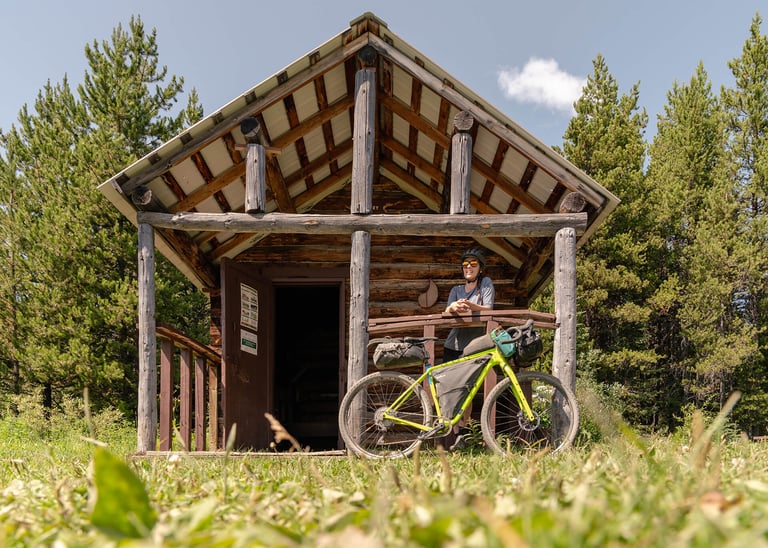 Bikepacker with a lime green bike beside a log cabin shelter among pines on a Canadian Rockies route.