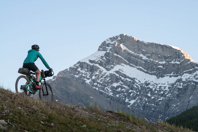 Bikepacker on a teal mountain bike climbs a grassy slope with a snow-streaked Rockies peak behind.