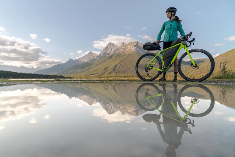 Rider with a lime green gravel bike and frame bag stands beside a reflective puddle with mountains behind.