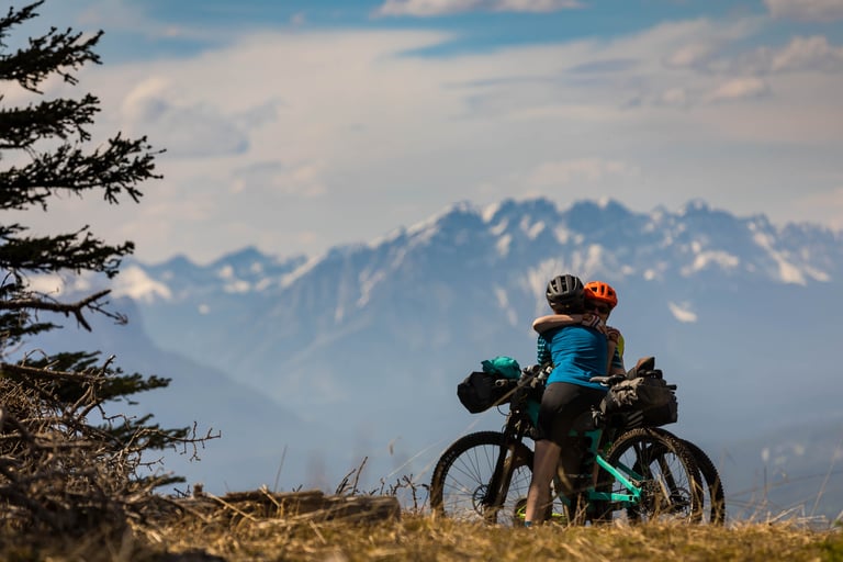 Two bikepackers embrace beside loaded bikes on a ridge with snowy Canadian Rockies peaks behind.