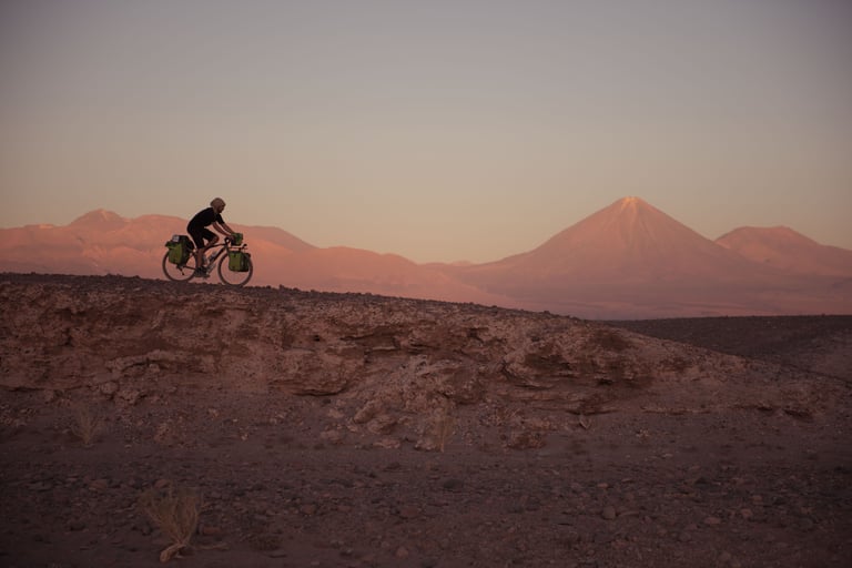 Touring cyclist with green panniers rides a desert ridge at sunset towards a conical volcano.