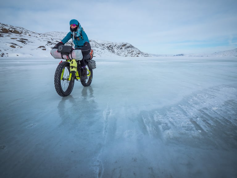 Fat bike rider with loaded bikepacking bags crosses a frozen lake below snowy hills.