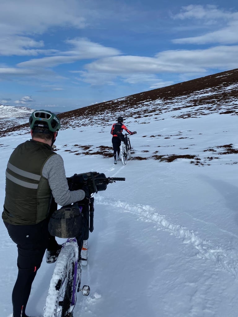 Two riders push loaded bikes through deep snow up a hillside on the Cairngorms bikepacking route.