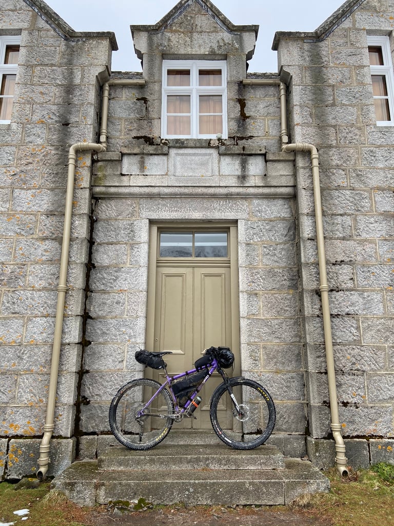Purple bikepacking bike with frame, bar and seat bags leans against the granite doorway of Glas-allt-Shiel bothy.