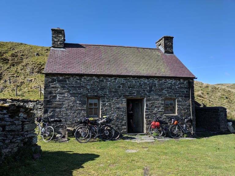 Bikepacking bikes leaning against the slate-roofed stone wall of Claerddu Bothy under a clear blue sky.