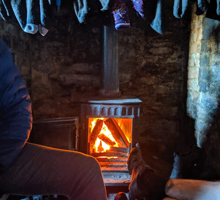 Log burner glowing inside a bothy, with socks hanging above it to dry and feet stretched towards the warmth.