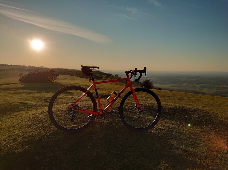 Red gravel bike with a saddle bag on a grassy hilltop at sunset, looking over open countryside during lockdown.
