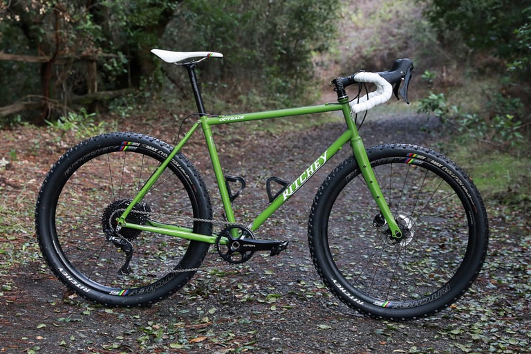 Green Ritchey Outback V2 gravel bike side-on on a leaf-strewn track, with white bar tape and knobbly tyres.