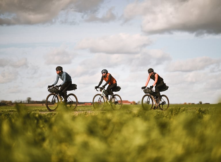 Three riders with loaded bikepacking bikes cross flat green farmland under a cloudy sky in Flanders, Belgium.