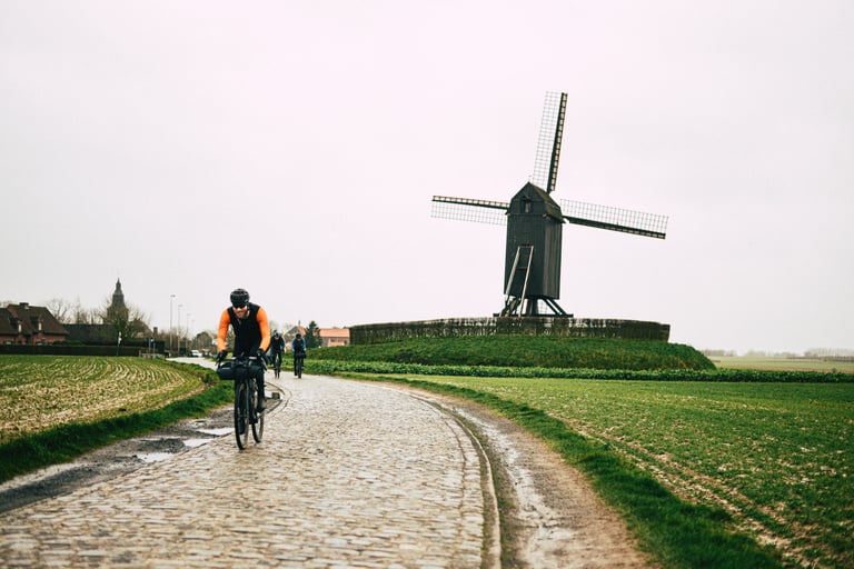 Rider on a wet cobbled lane past a black windmill in Flanders, with two more riders following in the rain.