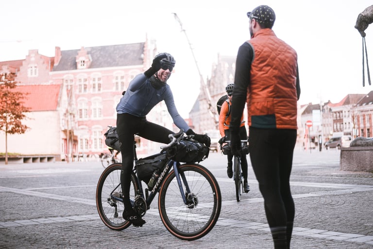 Smiling rider on a loaded Specialized gravel bike waves to a companion in a cobbled Belgian town square.