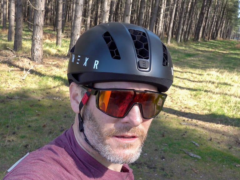 Rider wearing the HEXR helmet and orange-lens sunglasses in a pine forest, seen from the front.