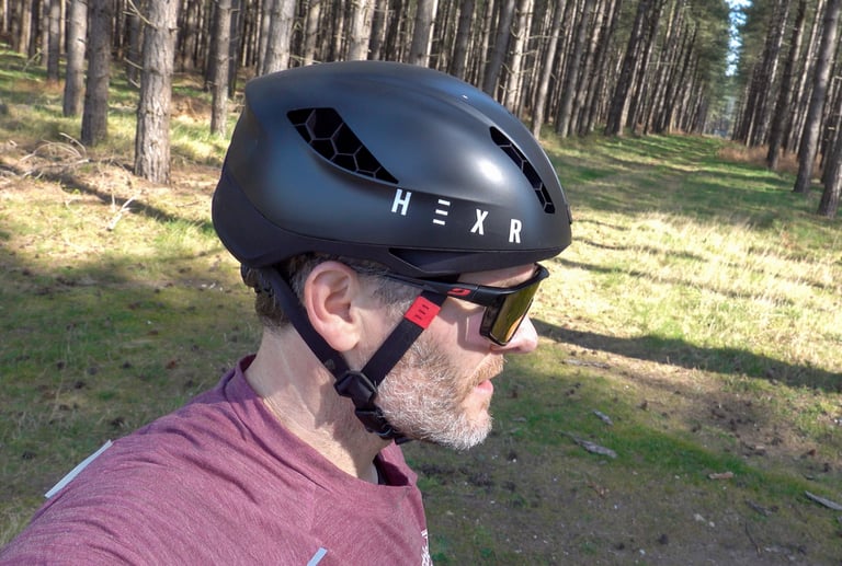 Side profile of a rider in the HEXR helmet in a pine forest, its deep side coverage sitting over the sunglasses arms.