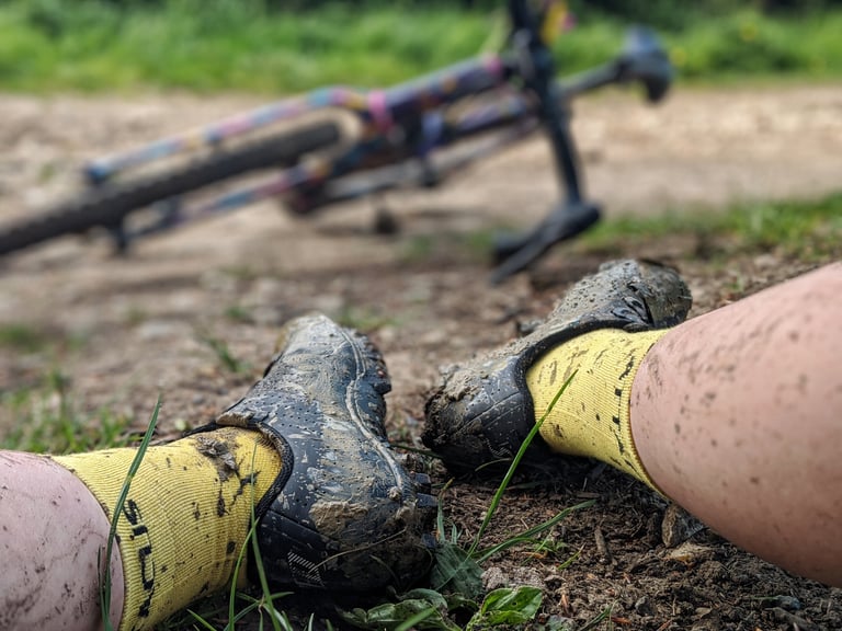 Muddy Specialized Recon 3.0 shoes and yellow socks on the grass after a ride, with a gravel bike lying behind.