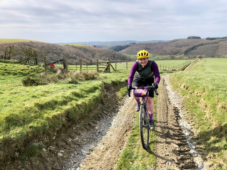 Rider on a gravel bike with a handlebar bag climbs a muddy farm track in rolling green hills.