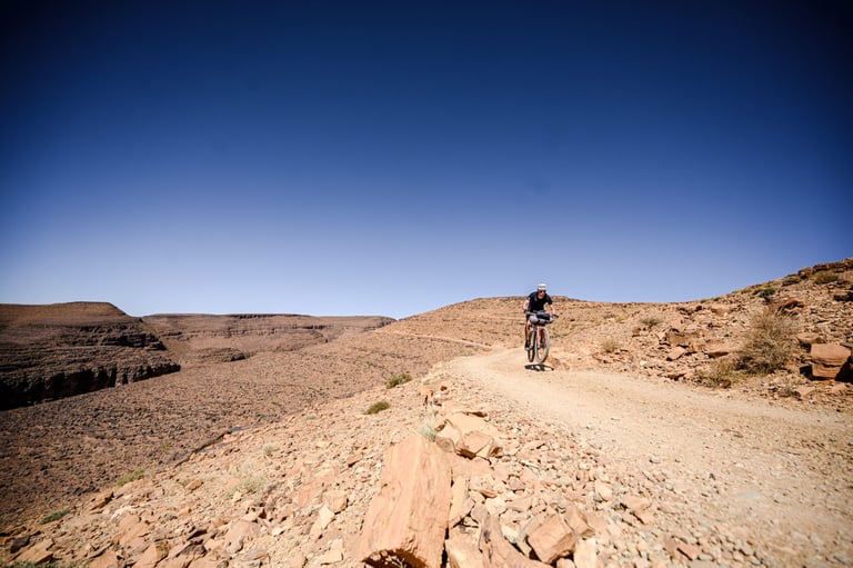 Atlas Mountain Race rider on a loaded bike follows a rocky desert track under a deep blue sky in Morocco.